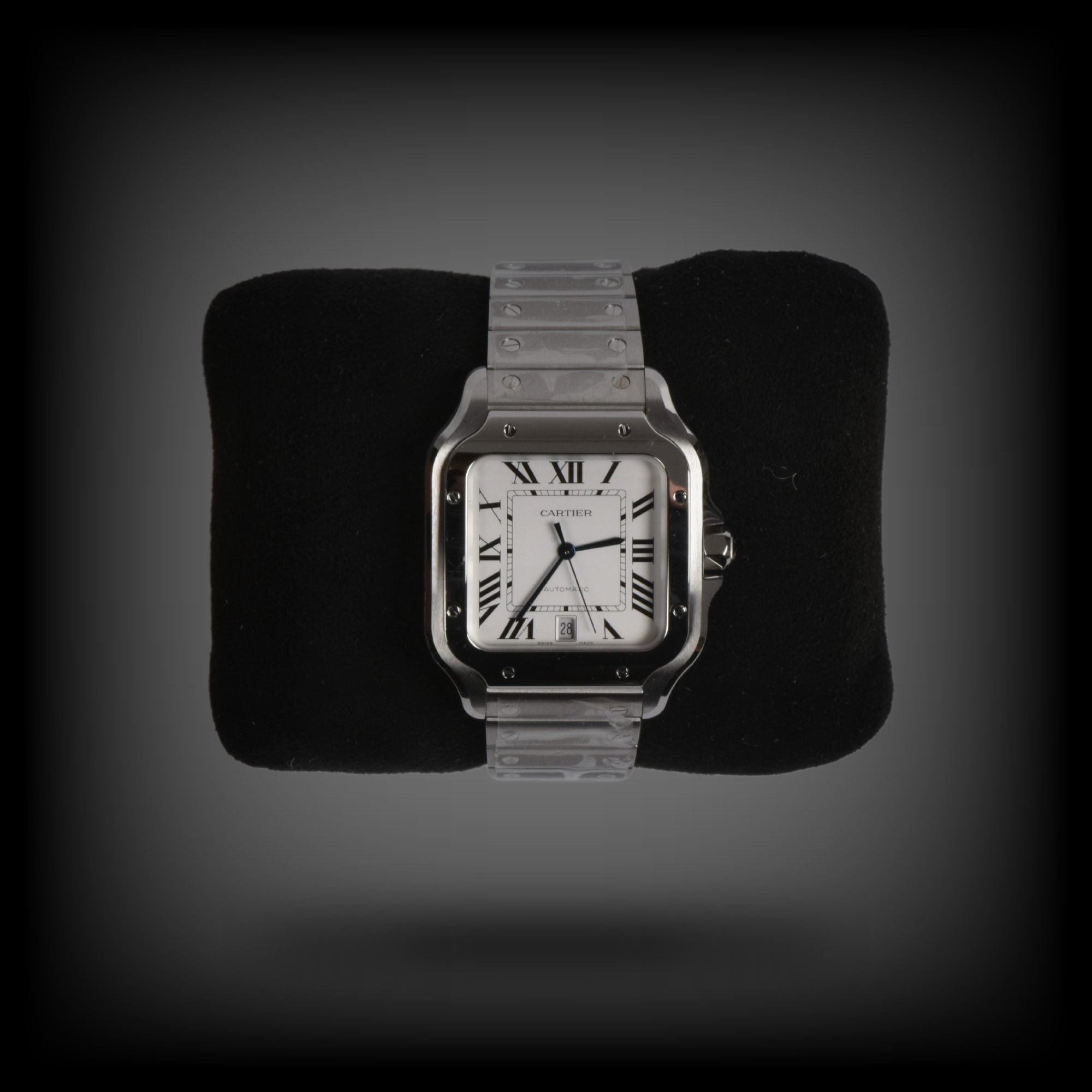 Eine silberne Armbanduhr mit eckigem Gehäuse liegt auf einem schwarzen Kissen. Die Uhr hat ein weißes Zifferblatt, schwarze römische Ziffern und drei Zeiger. Auf dem Zifferblatt steht der Markenname „Cartier“. Das Armband besteht aus silbernem Metall mit sichtbaren Schraubendetails. Die Lünette wirkt poliert, das Datumsfenster befindet sich bei sechs Uhr. Der Hintergrund ist einfarbig und dunkel gehalten, wodurch die Uhr besonders hervorgehoben wird.