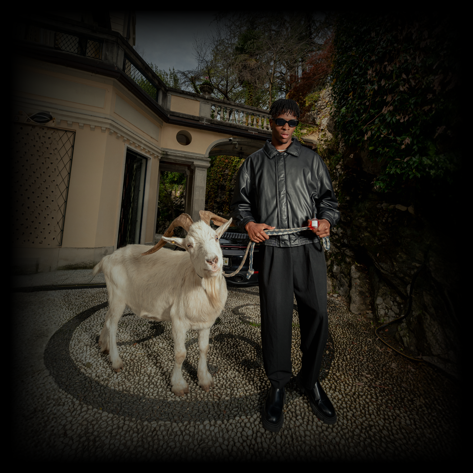 Ein Mann mit schwarzer Sonnenbrille, schwarzer Lederjacke und dunkler Hose hält eine weiße Ziege an einer Leine. Im Hintergrund steht ein elegantes Gebäude mit cremefarbener Fassade, daneben sind ein Autogrill und abwechslungsreiche grüne Pflanzen zu sehen. Der Boden ist mit einem Muster aus kleinen Steinen in Kreisen gestaltet. Die Atmosphäre wirkt luxuriös und stilvoll.